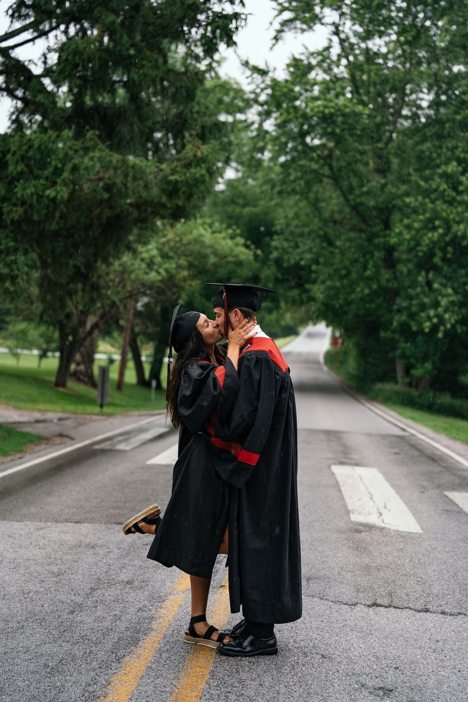 Two graduates in black and red gowns share a romantic kiss on a tree-lined road on graduation day.