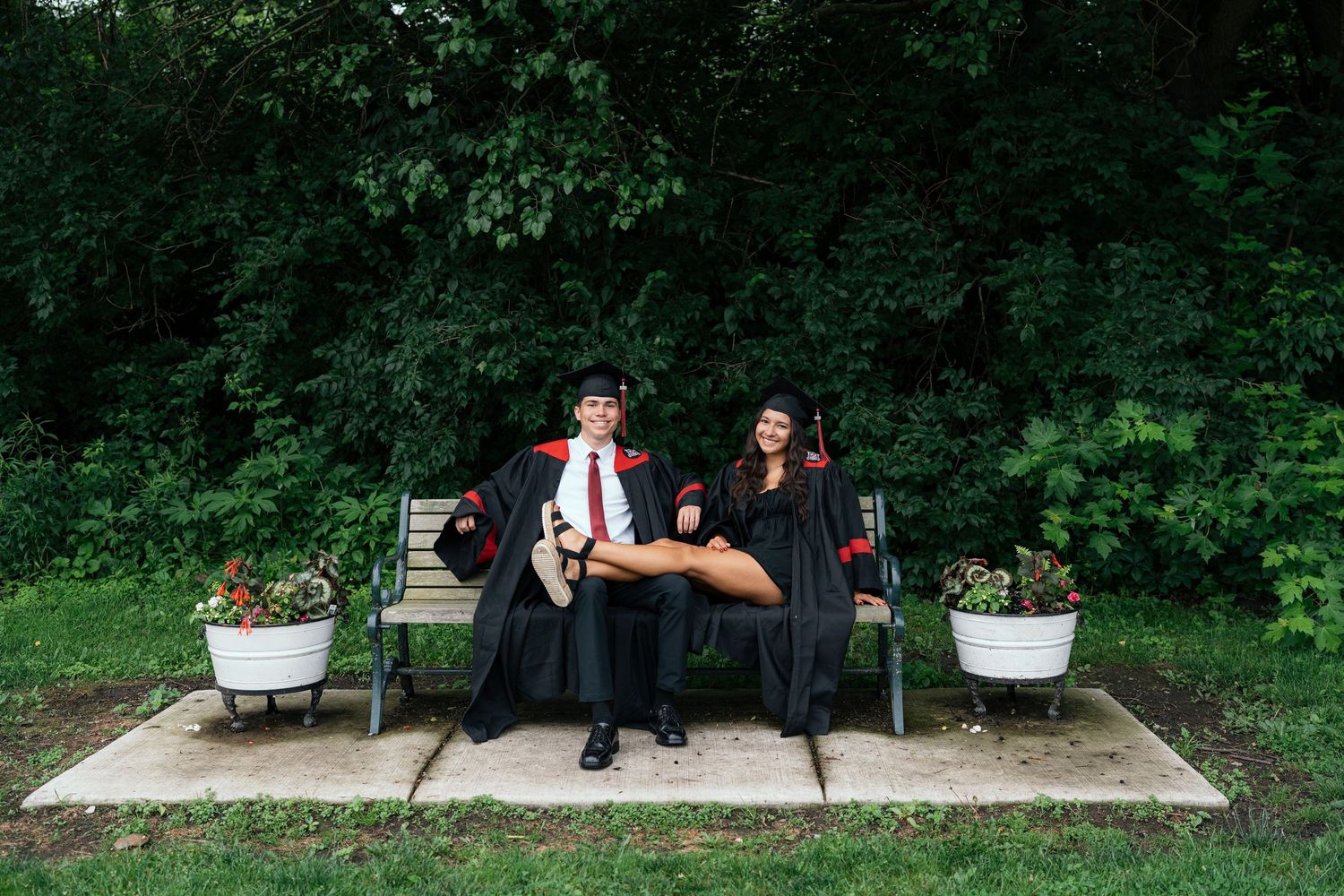 Two graduates in black robes sit on a bench outdoors, surrounded by lush greenery and flower pots.