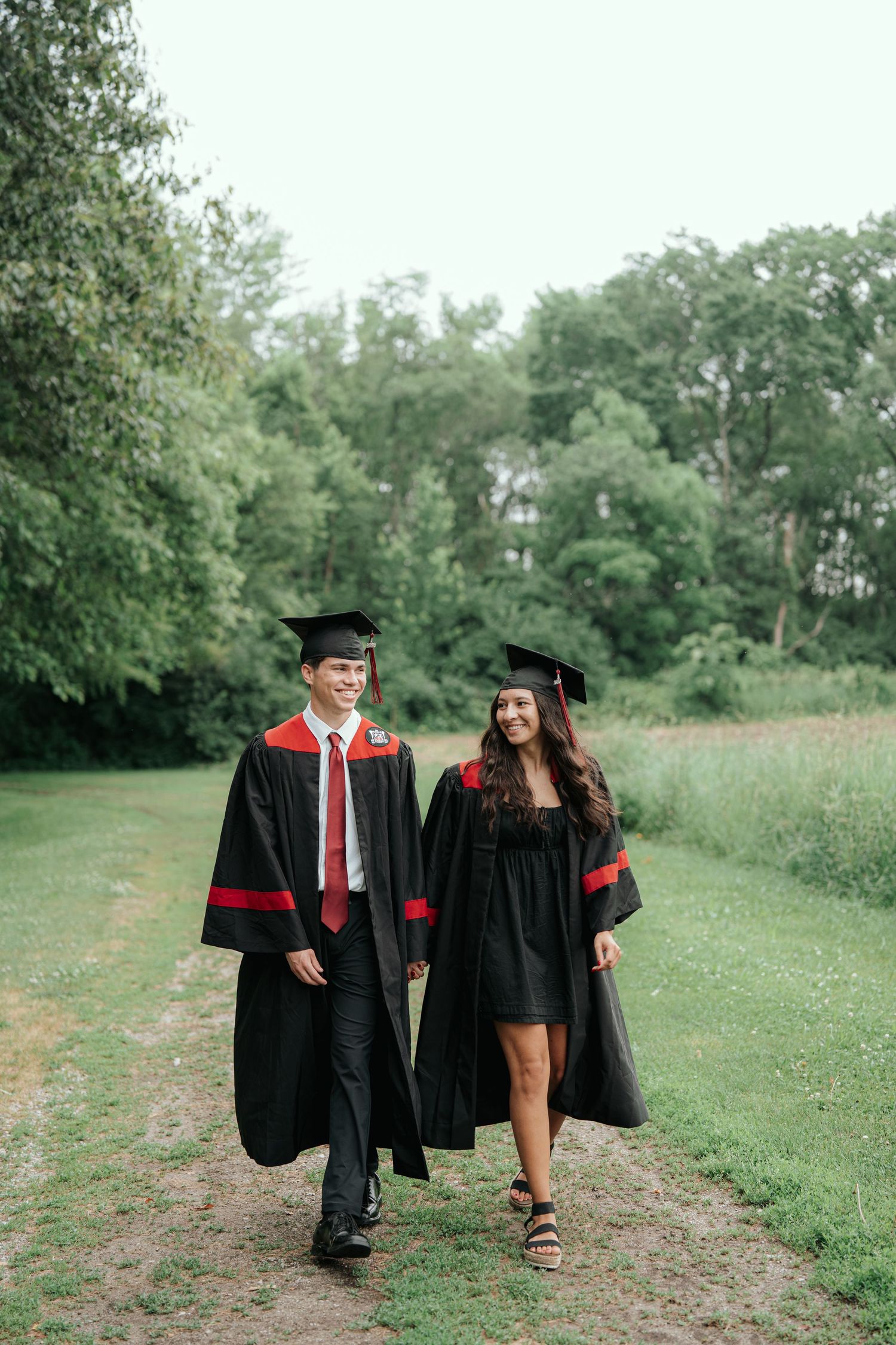 Two graduates in black caps and gowns walk together on a garden path, smiling and celebrating their achievement.
