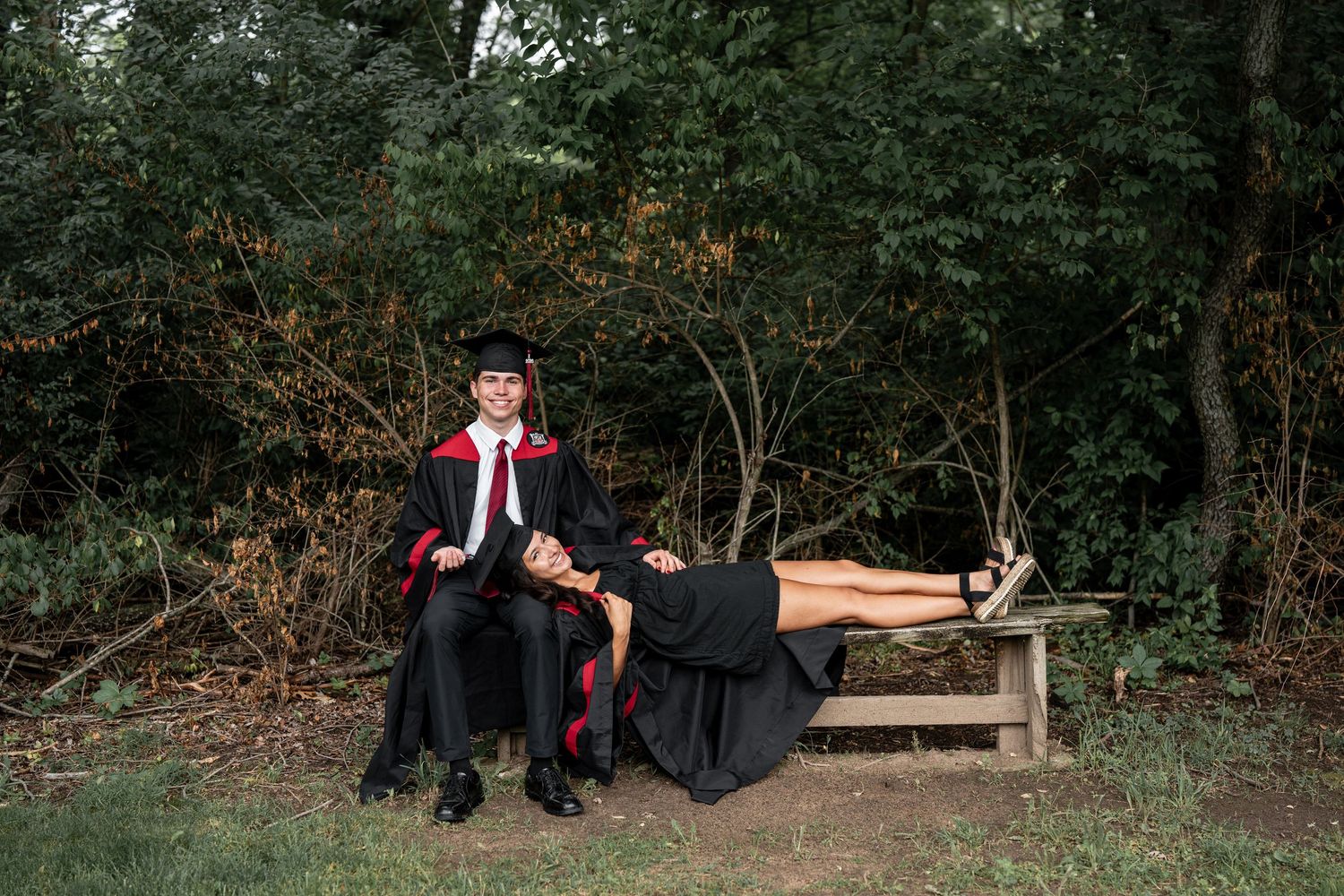 Graduate in cap and gown sitting on a bench outdoors with a companion lying across, surrounded by lush green trees.