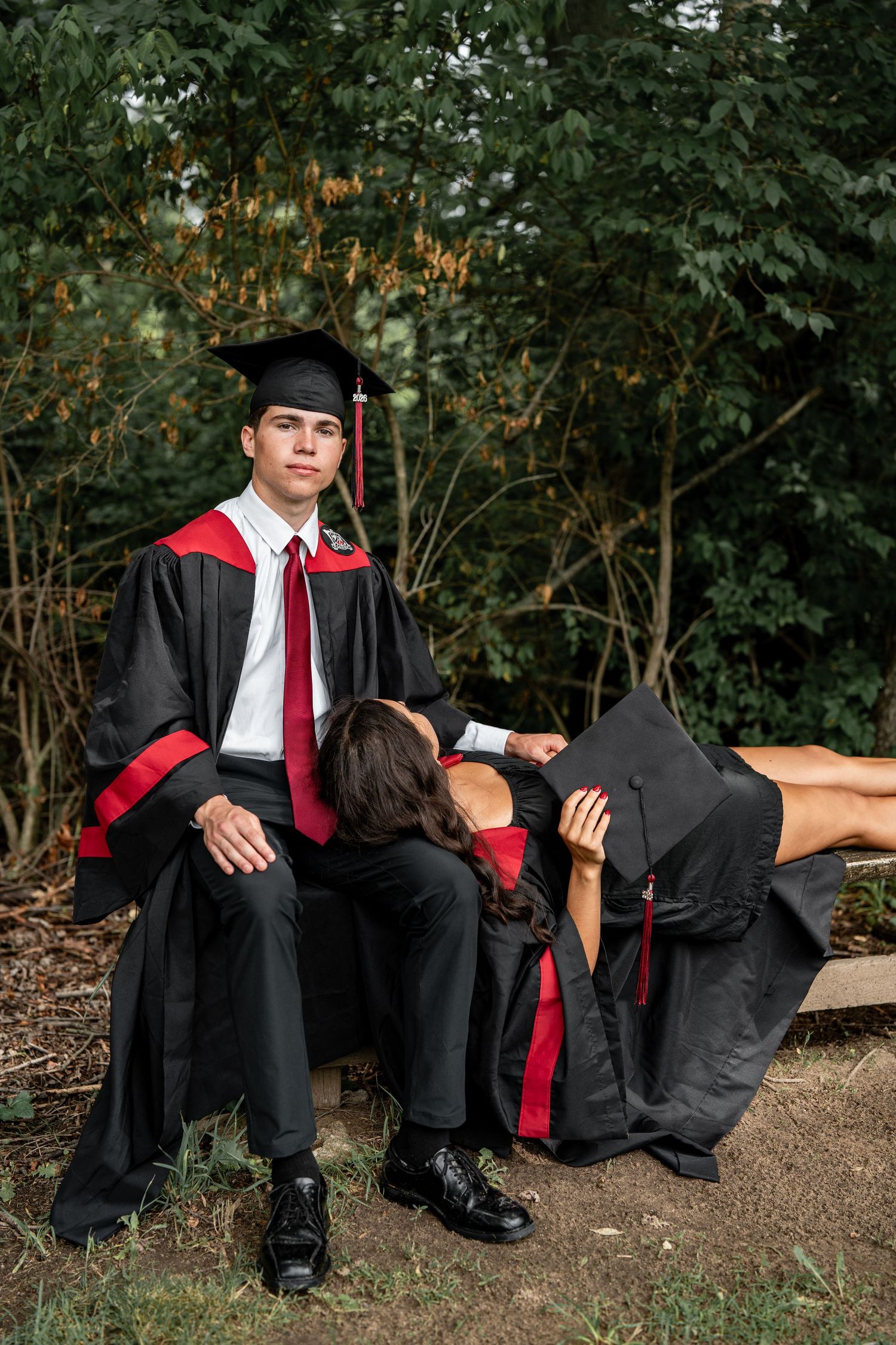 Male graduate in cap and gown sitting outdoors while female graduate rests beside him.