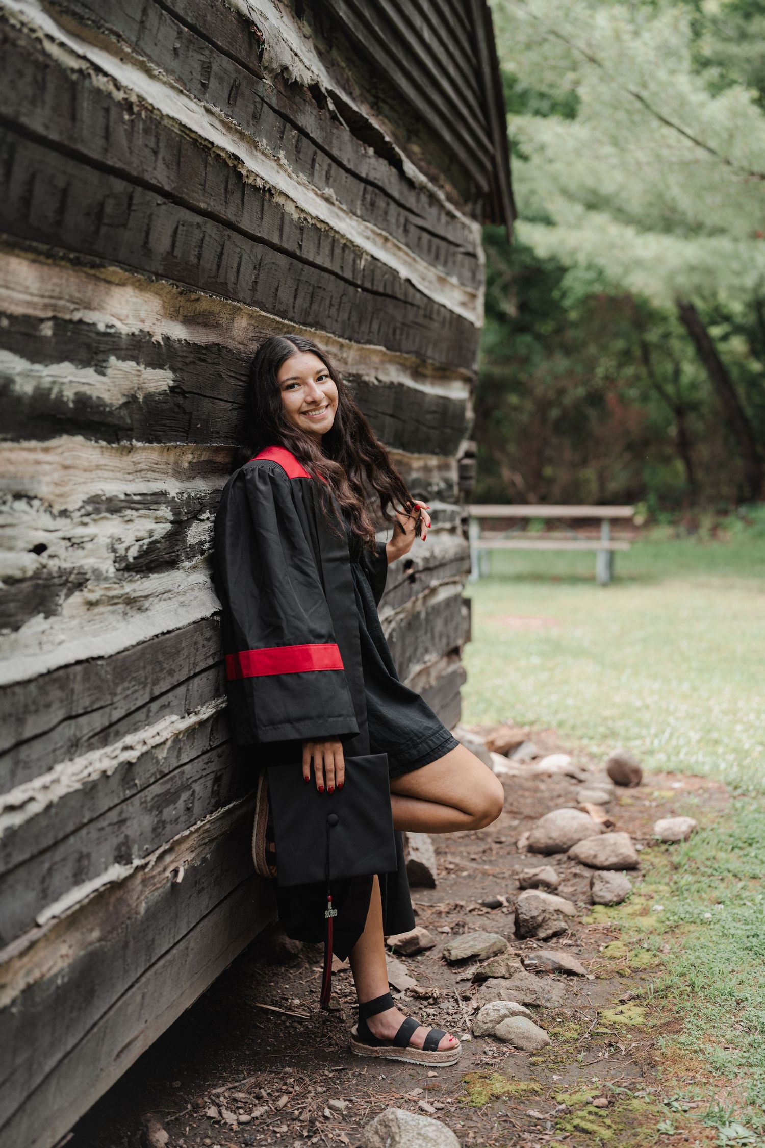Young woman in black and red graduation gown leaning against rustic log cabin wall, smiling outdoors.