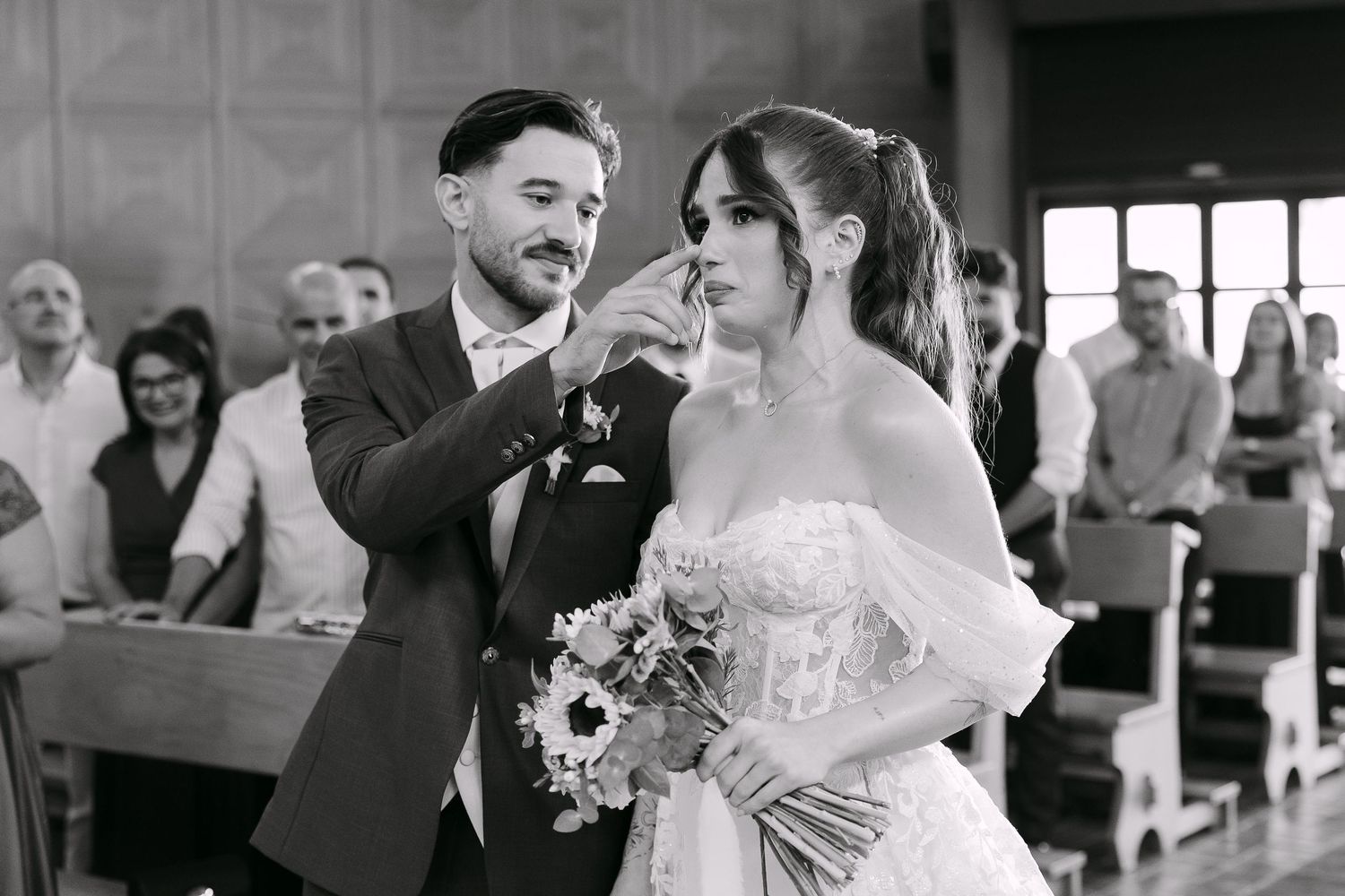 Groom gently wipes bride's tear during emotional wedding ceremony in church, bride holding bouquet.