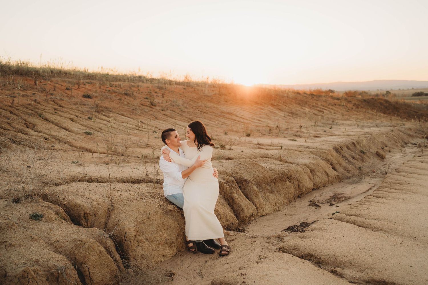 Couple sitting together on sandy terrain at sunset in a romantic outdoor portrait.