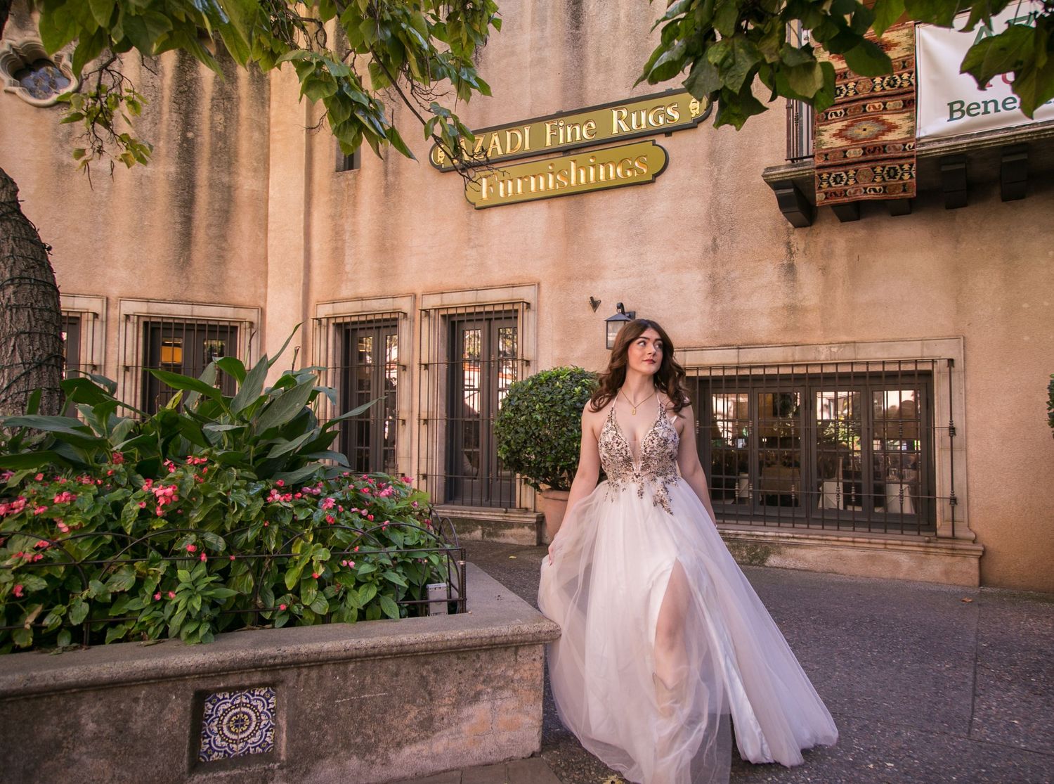 Woman in elegant floral ballgown posing outside a rustic building with lush greenery and signage.