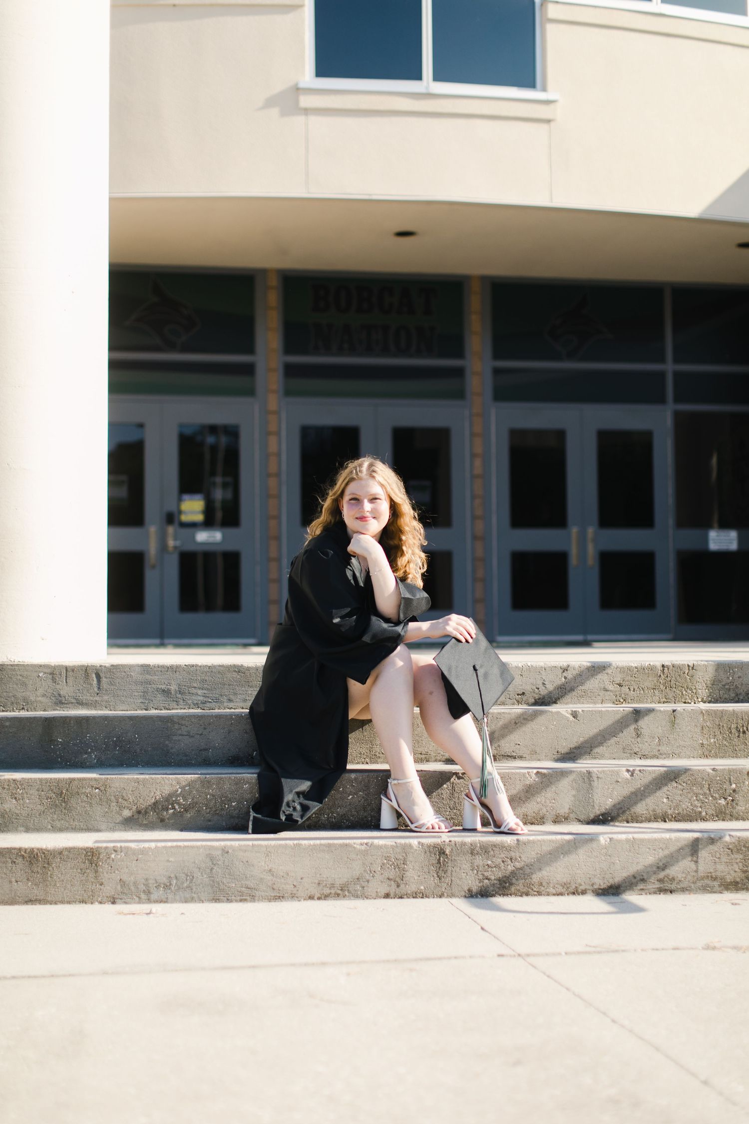 A person in dark clothing sits on concrete steps outside a school with large windows in a casual outdoor portrait setting at Bluffton High.