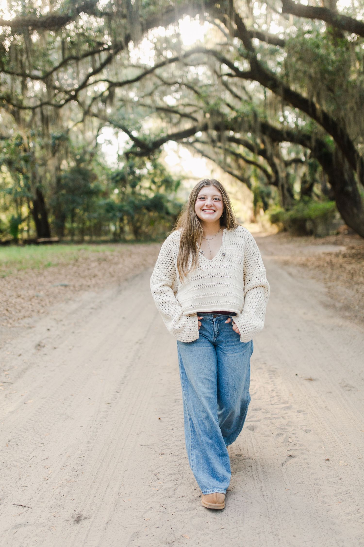 A person walks down a dirt road lined with Spanish moss-draped oak trees wearing a cream sweater and blue jeans for Beaufort senior session.