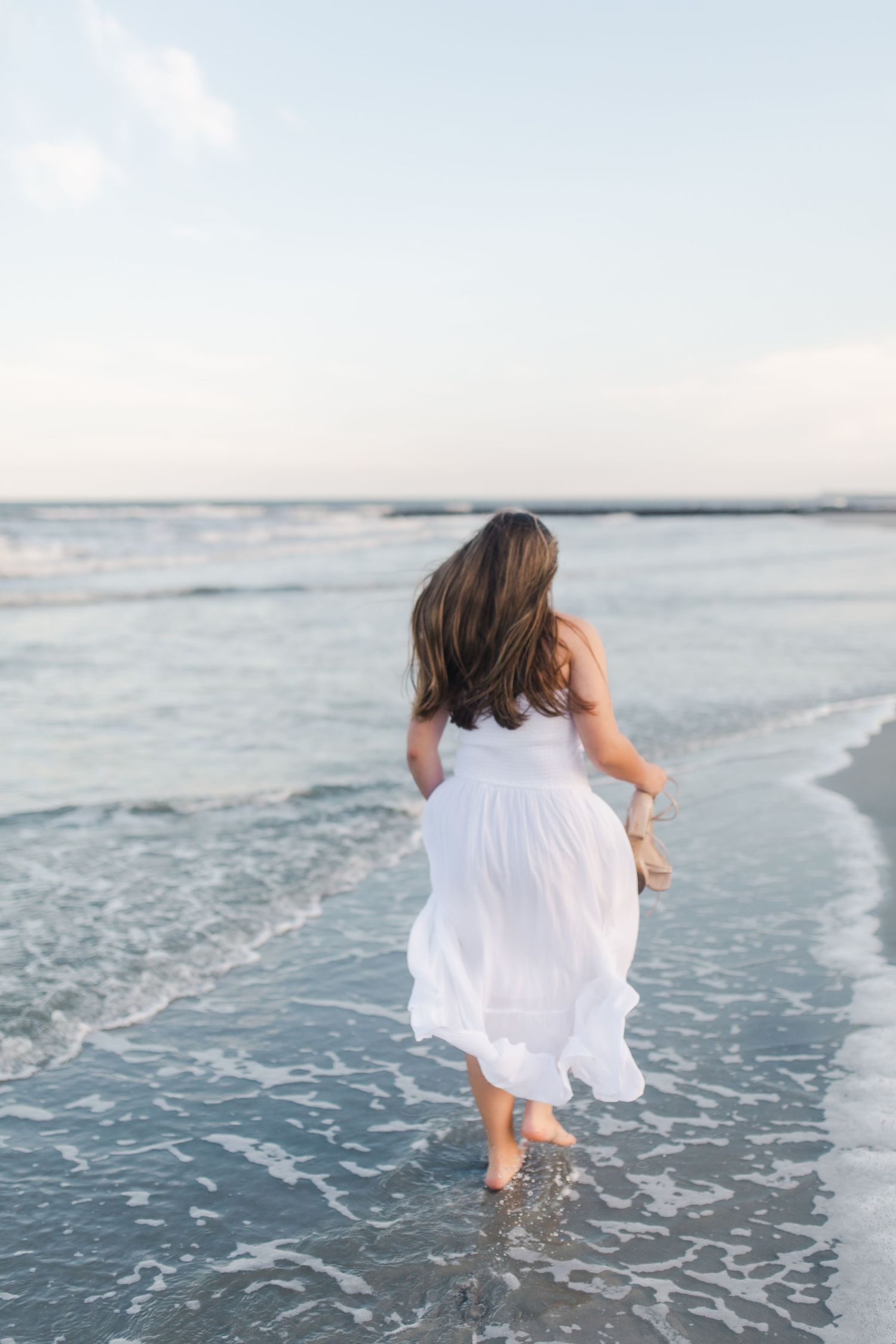 A person in a flowing white dress runs joyfully along the ocean shoreline at sunset.