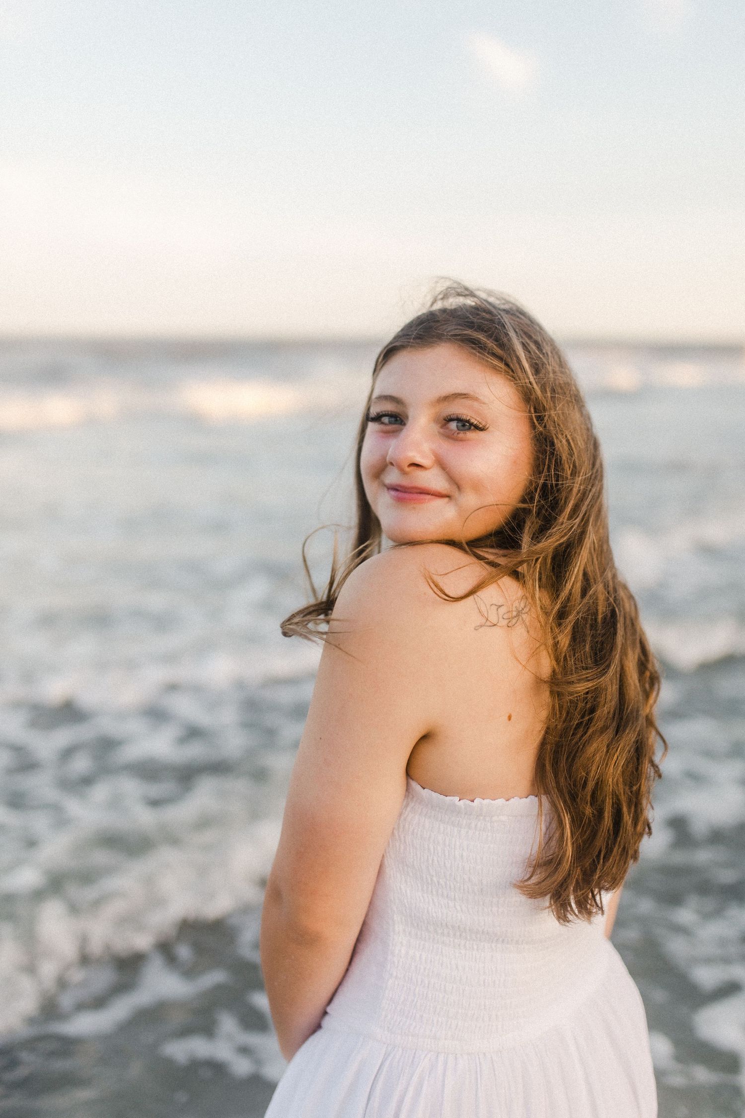 A person in a white dress poses joyfully by the ocean during sunset, with their hair blowing in the sea breeze at Hunting Island beach.