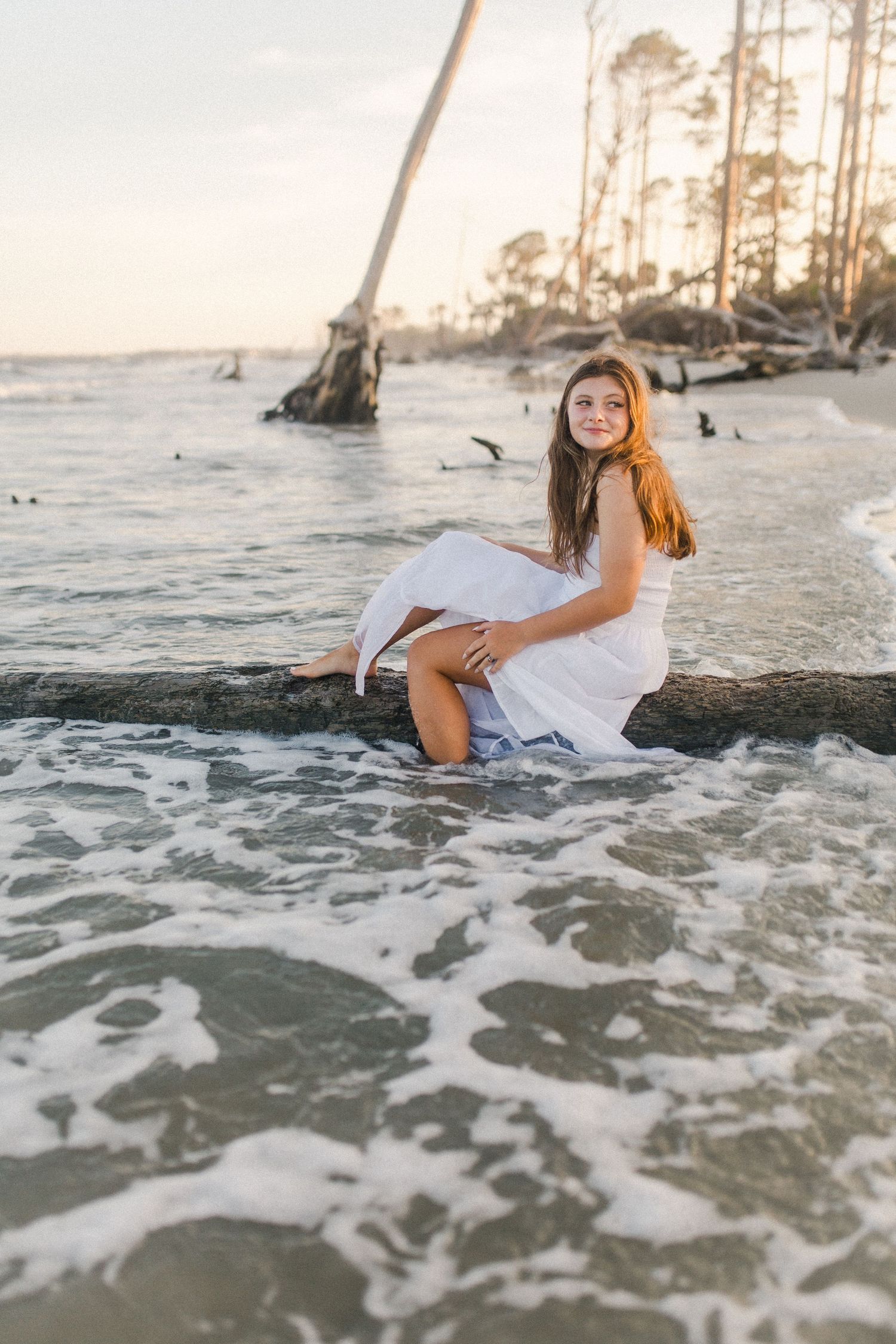 A senior session of a girl in a white dress lying on a log at the beach during sunset as waves crash around them on Hunting Island beach.