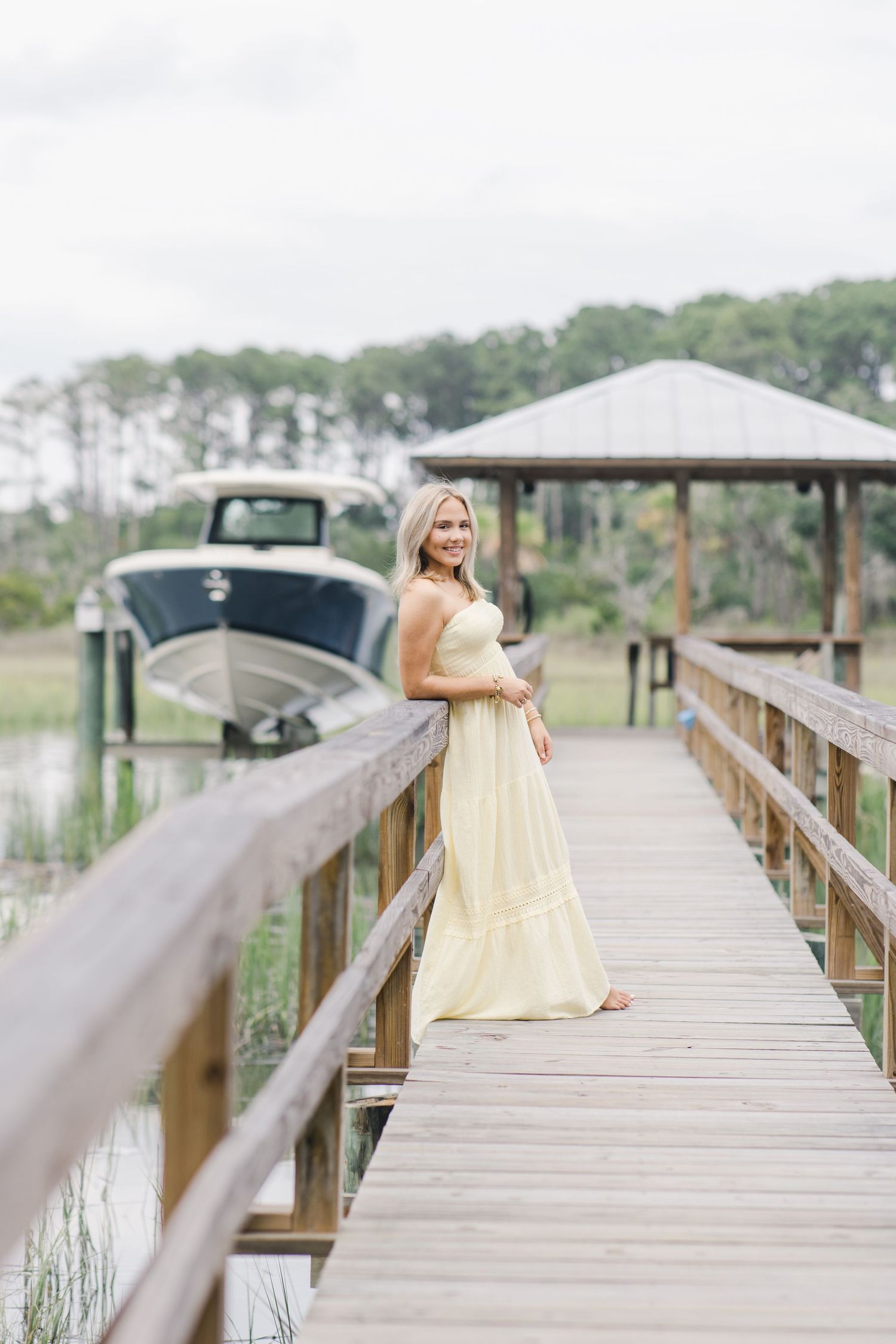 A person in a yellow dress poses on a wooden lowcountry boardwalk over marshland on a cloudy day for senior photos.