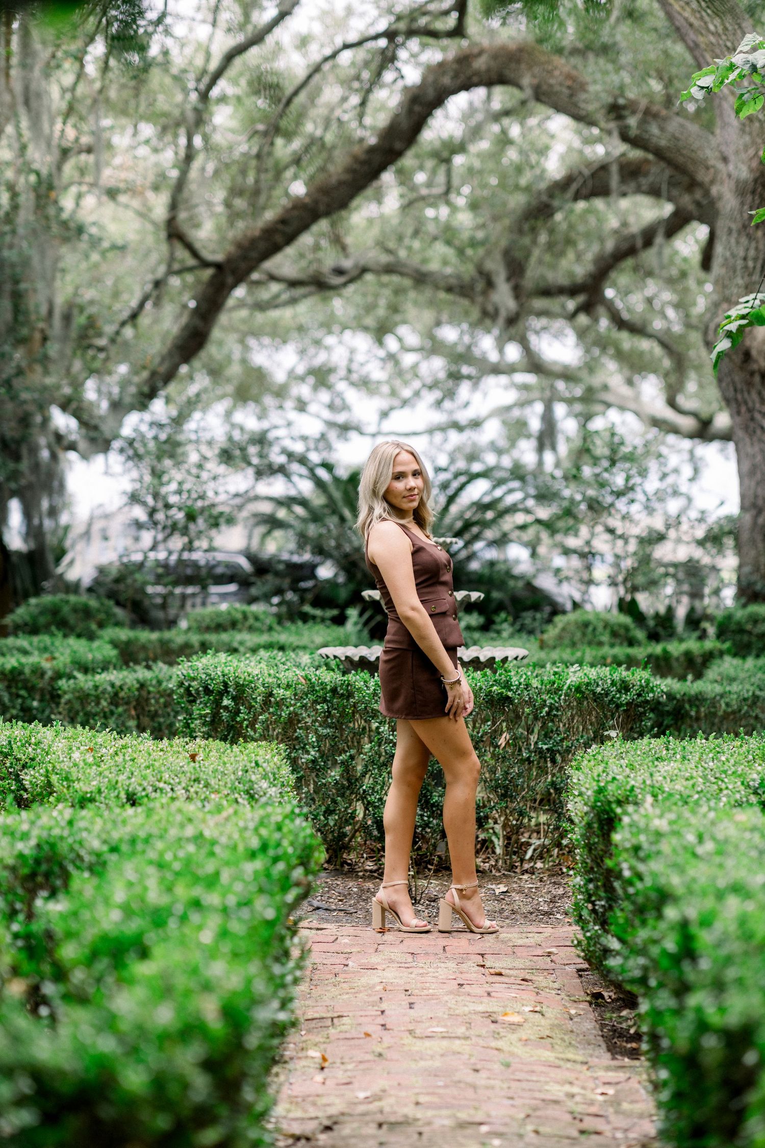 A person in brown clothing walks along a brick path surrounded by lush green hedges and trees in a garden setting.