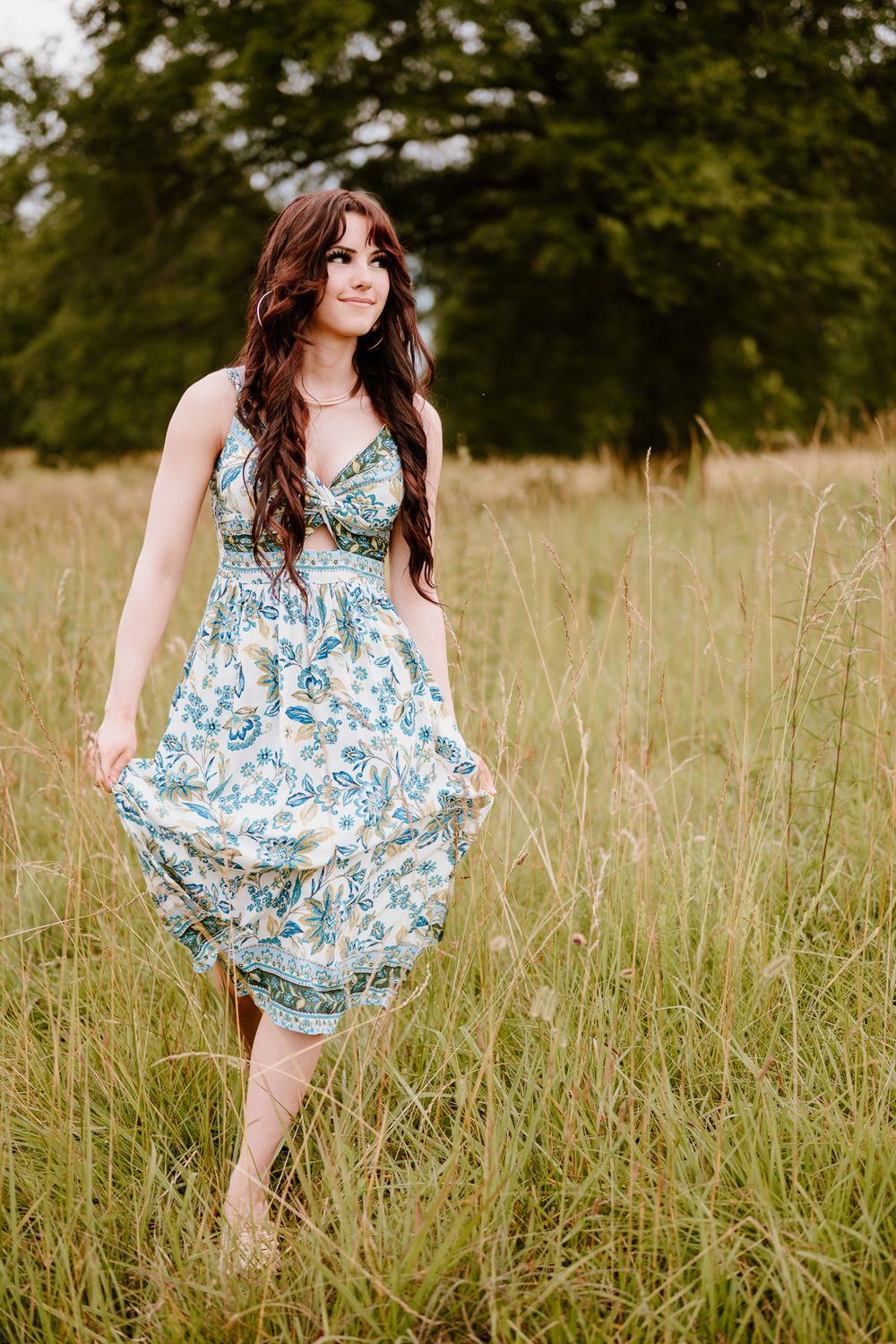 A woman in a flowing blue floral sundress stands barefoot in a summer meadow with tall grass and trees in the background.