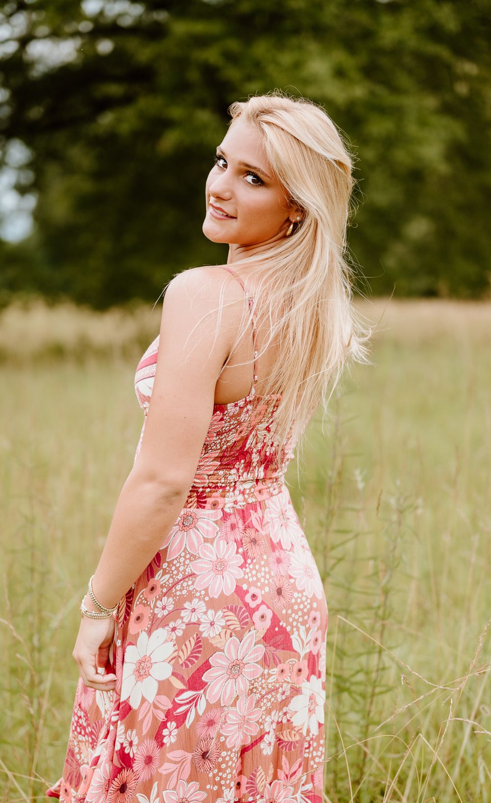 A person poses in a pink floral sundress while standing among tall grass in a natural outdoor setting.