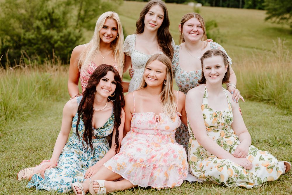A group in floral summer dresses pose together on a grassy hillside during a warm afternoon photoshoot.