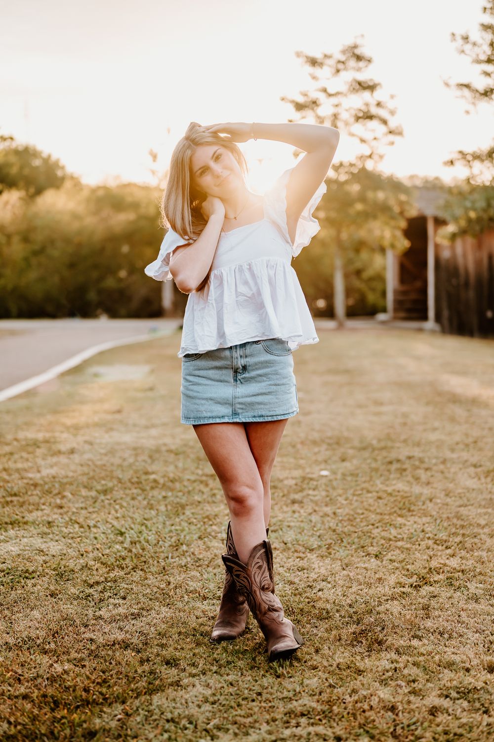 Person wearing brown cowboy boots and denim shorts stands in a grassy field at sunset.