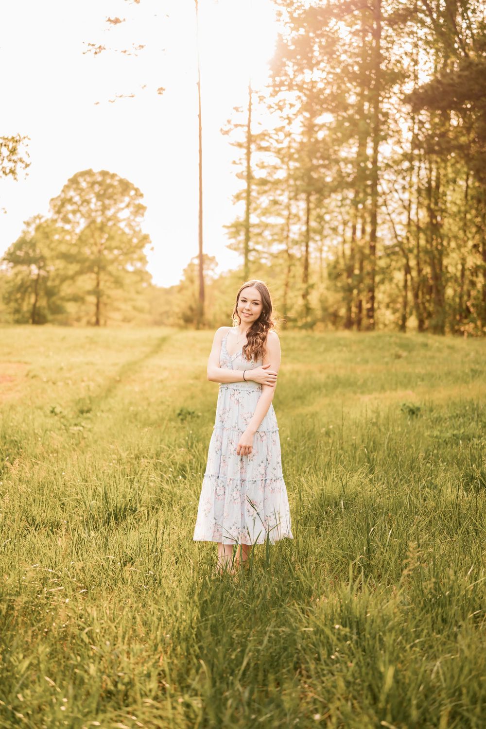 A High School Senior wearing a white lace dress stands in a sunlit meadow at golden hour with tall trees in the background.