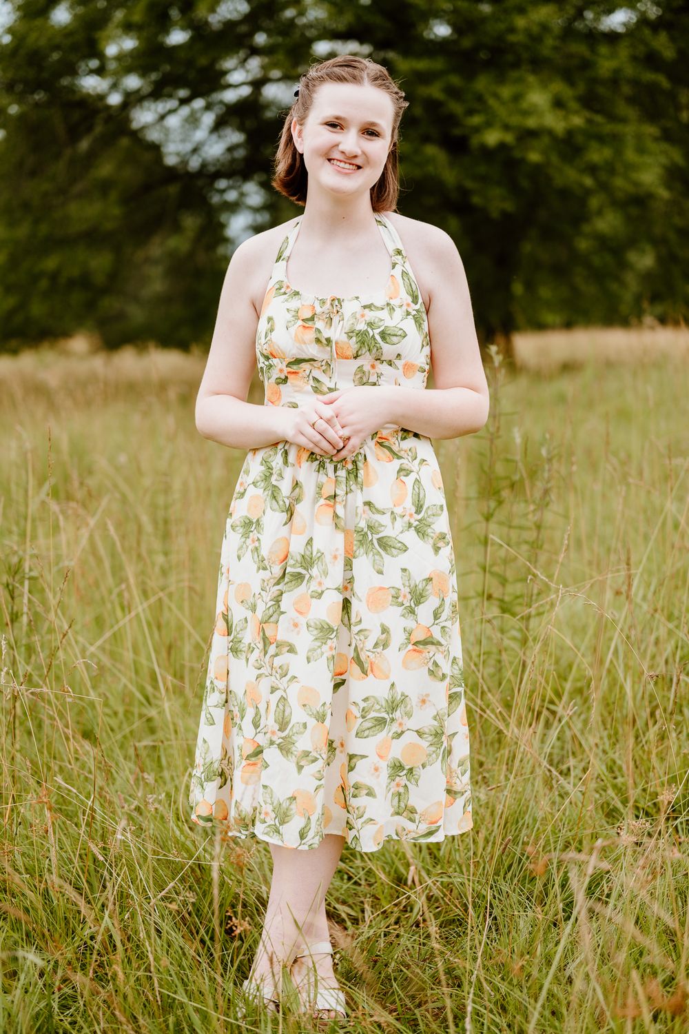 A young girl in a floral summer dress stands playfully in a grassy meadow on a sunny day.