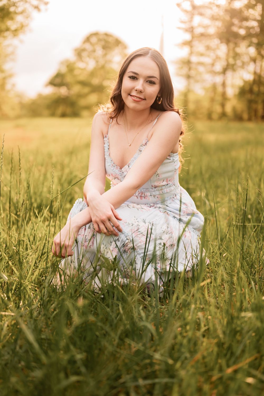 A High School Senior in a white floral dress sits in tall grass at sunset with warm golden light filtering through trees.