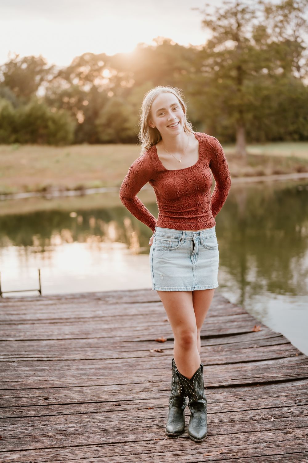 A person in a red top and denim skirt poses playfully on a wooden dock by a lake at sunset.