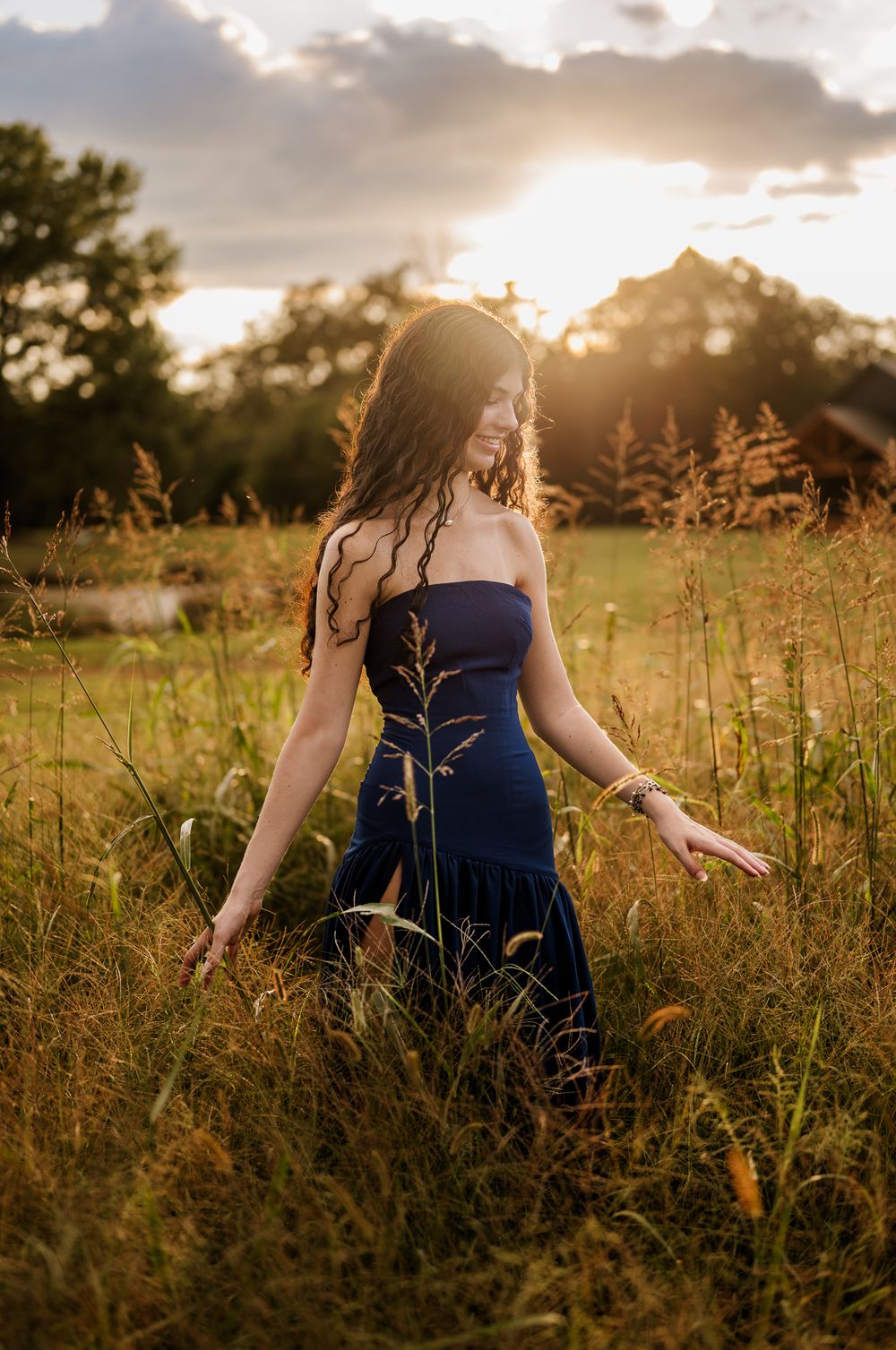 A person in a navy blue dress stands in a golden field at sunset with tall grass swaying in the breeze.