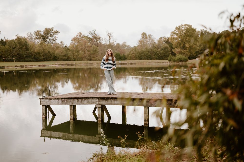 A long wooden pier extends across a tranquil lake surrounded by autumn trees reflecting in the calm water.
