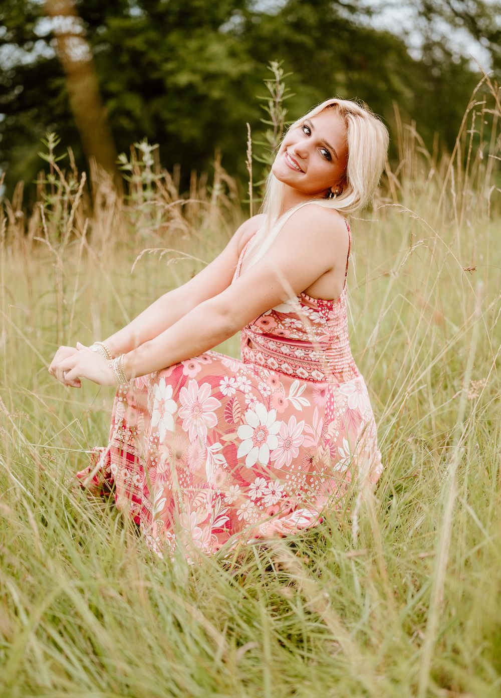 A person in a floral pink sundress twirls playfully in a field of tall summer grass on a sunny day.