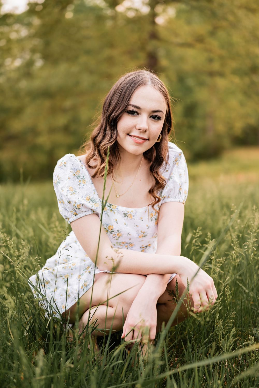 A High School Senior in a floral white dress crouches in a sunlit grassy meadow during golden hour.