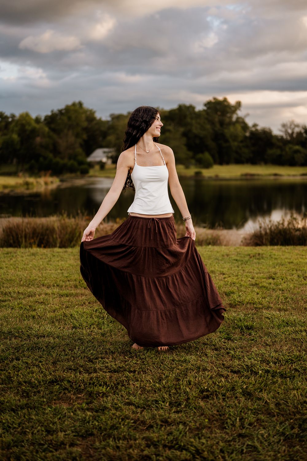 A person in a white top and brown skirt twirls gracefully in a field by a lake during golden hour.