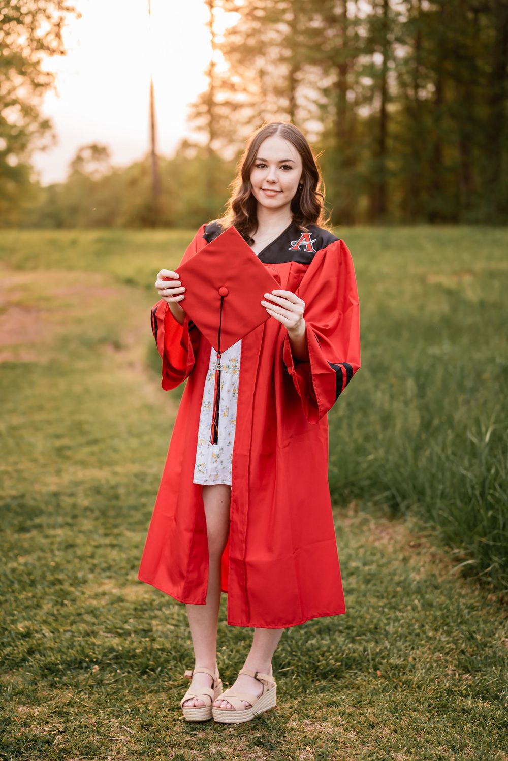 High School Senior standing in red cap and gown holds up diploma during golden hour sunset in outdoor setting.