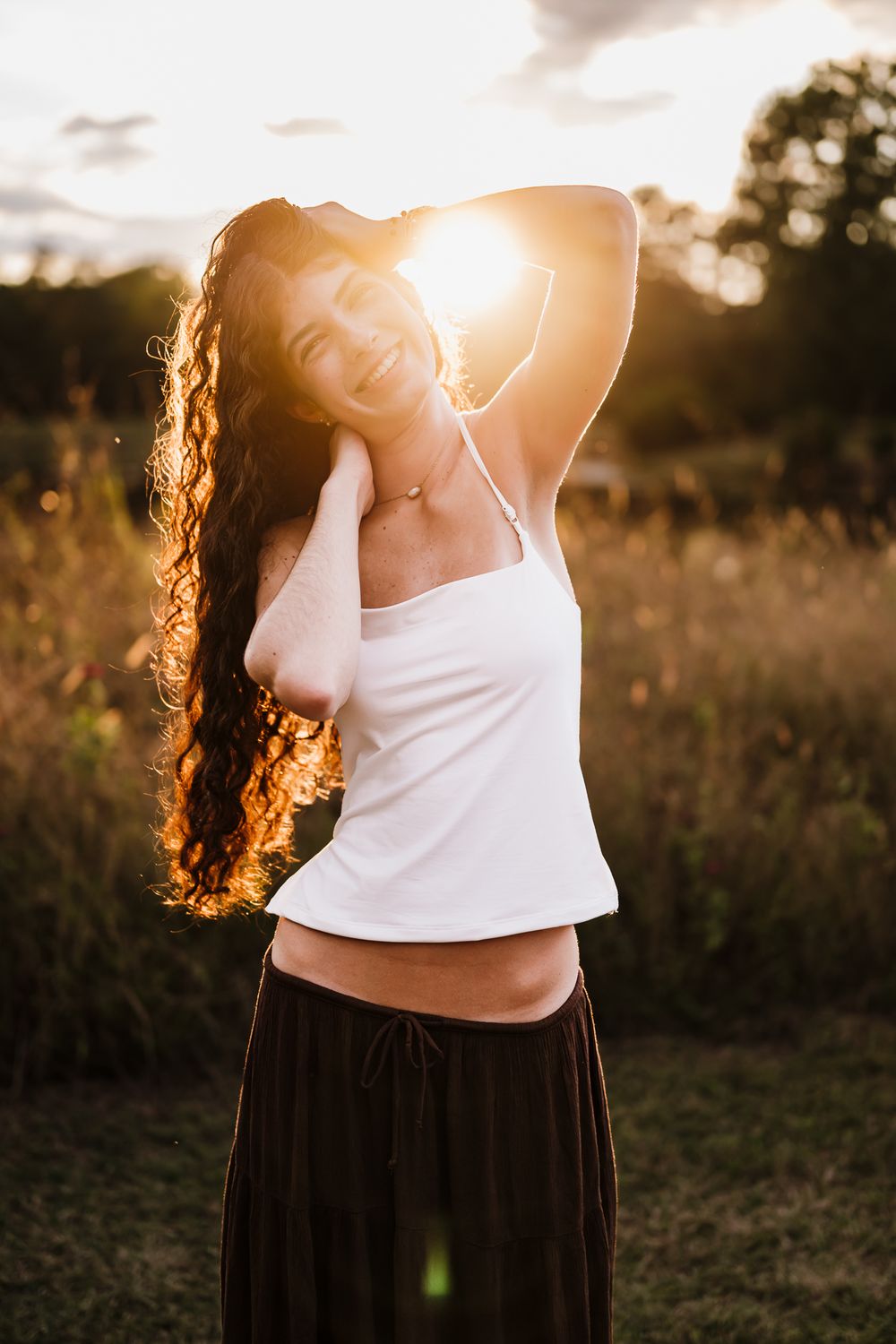 A backlit silhouette in a white tank top and dark skirt poses gracefully in a golden sunset field.
