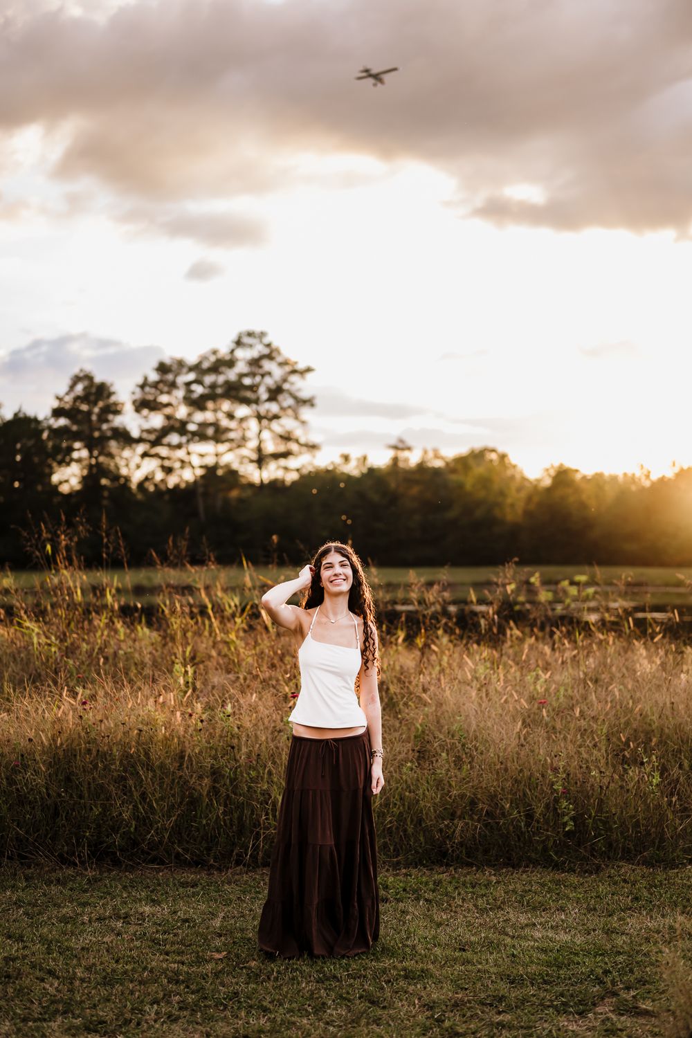 Silhouette of a person in a flowing skirt standing in a golden field at sunset with trees in the background.