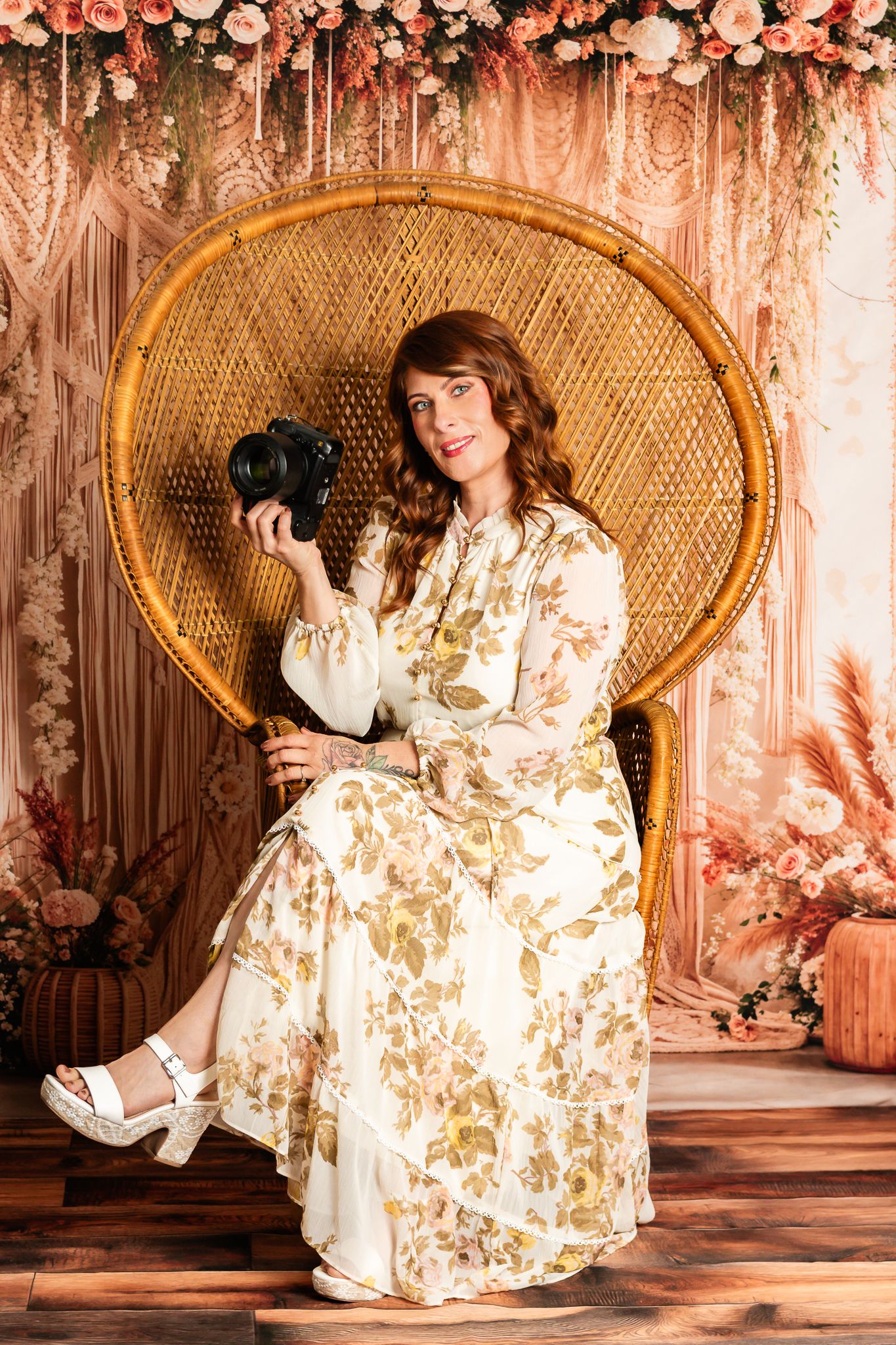 Professional photographer in vintage floral dress sits on peacock chair with camera against romantic pink backdrop.
