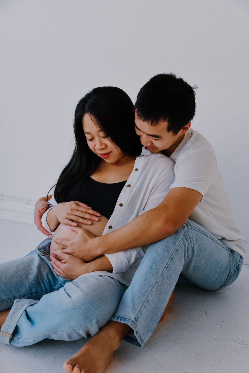 Expectant couple in casual wear sitting on floor during maternity shoot.