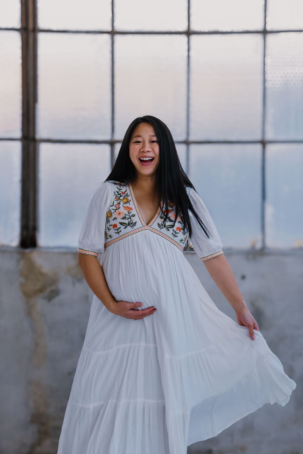 Woman twirls in white flowy dress against industrial windows.