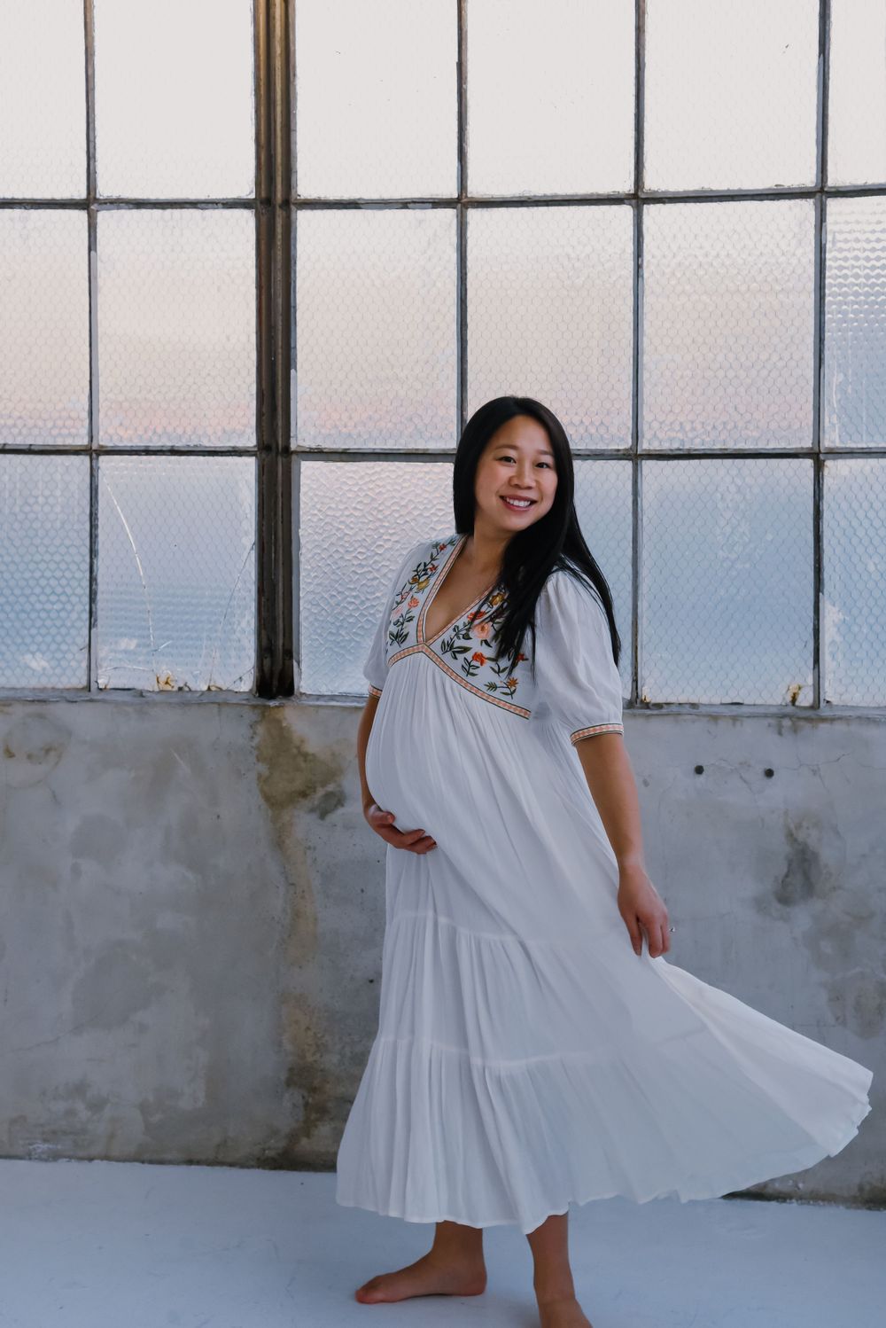 Woman dances playfully in white dress near large windows.