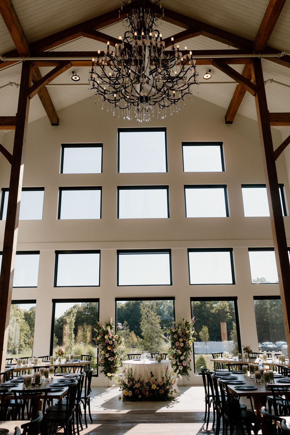 Elegant wedding venue interior with exposed wooden beams, chandeliers, and large windows filling the space with natural light.