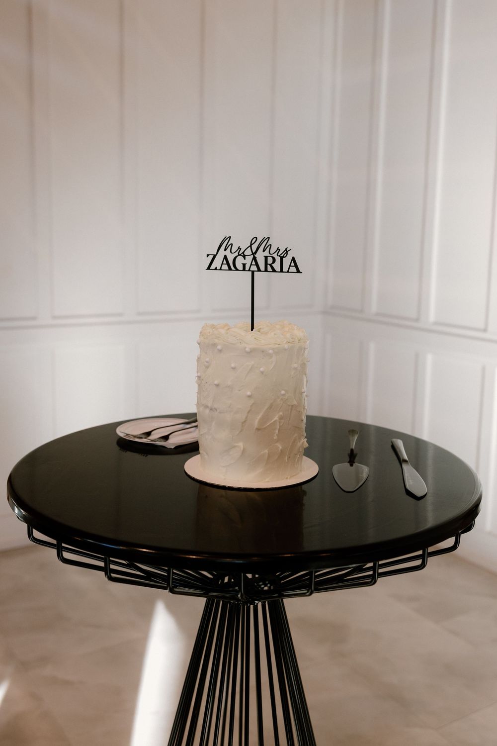 Modern black wire frame pedestal table with white frosted cake featuring a simple topper displayed in elegant setting.