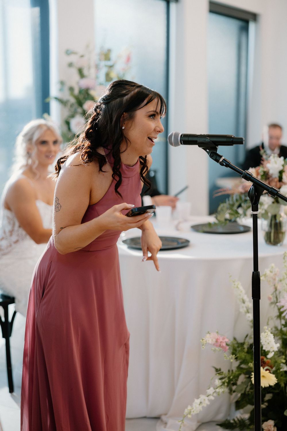 A bridesmaid in rose delivers an emotional wedding speech at a reception with decorative floral arrangements.