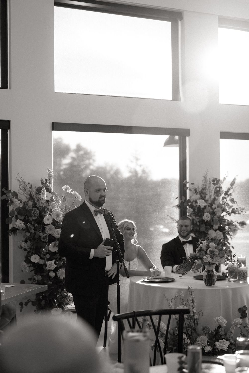 A formal wedding reception speech is given in an elegant room with large windows and floral arrangements.