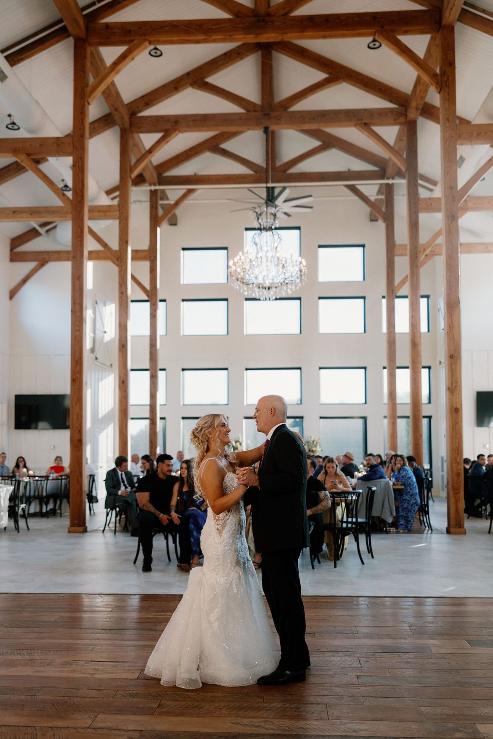 Two figures share a meaningful slow dance in a rustic venue with exposed wooden beams and tall windows.