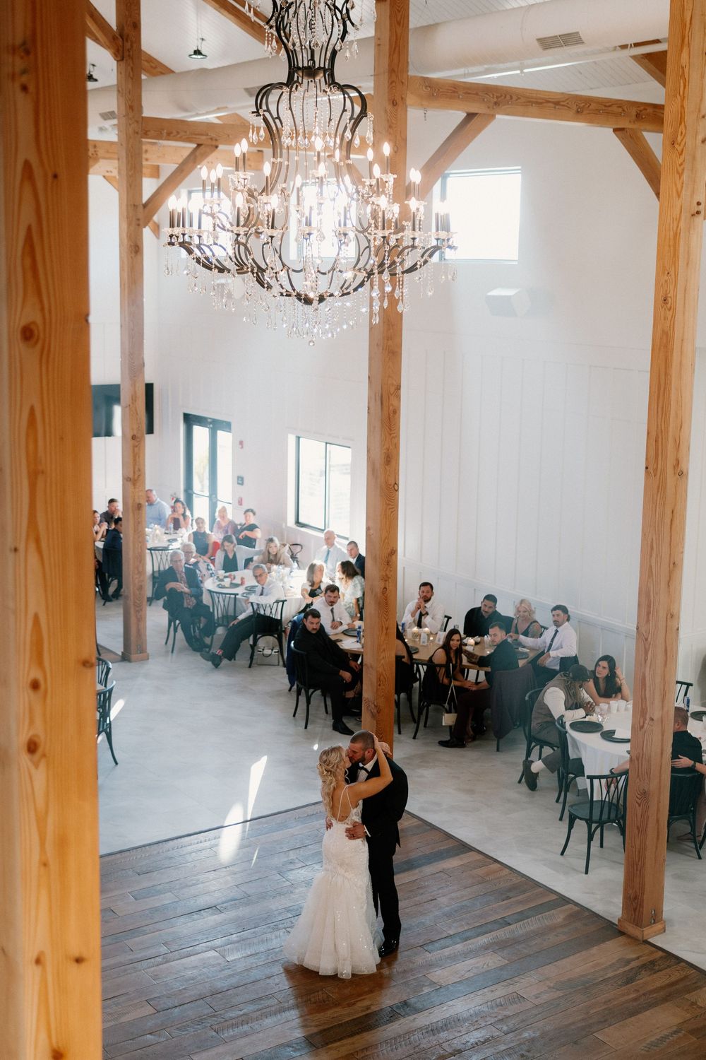 Elegant wedding reception venue featuring exposed wooden beams, crystal chandeliers and white decor with guests seated at tables.