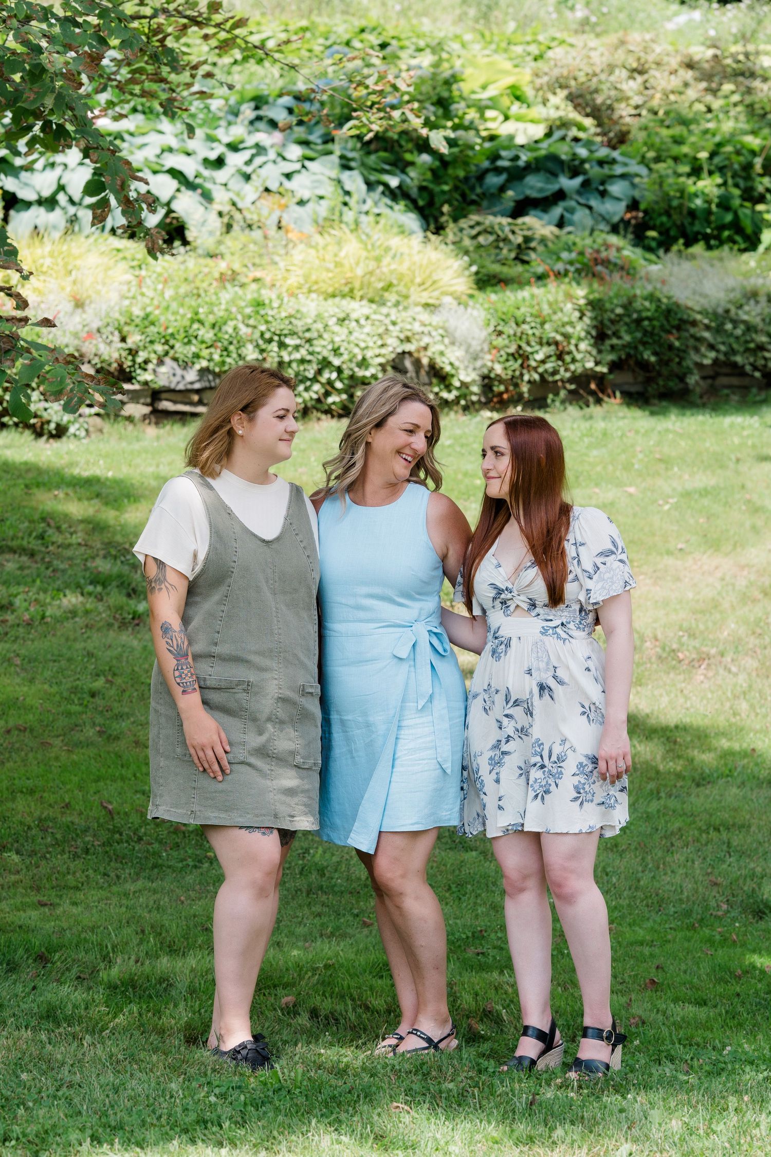 Three women smile and pose together in a lush green garden wearing casual summer dresses at Dartmouth Commons.