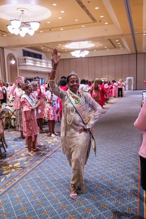 A Golden Soror of Alpha Kappa Alpha Sorority, Incorporated walks down the aisle while waving her hand as everyone claps for her.