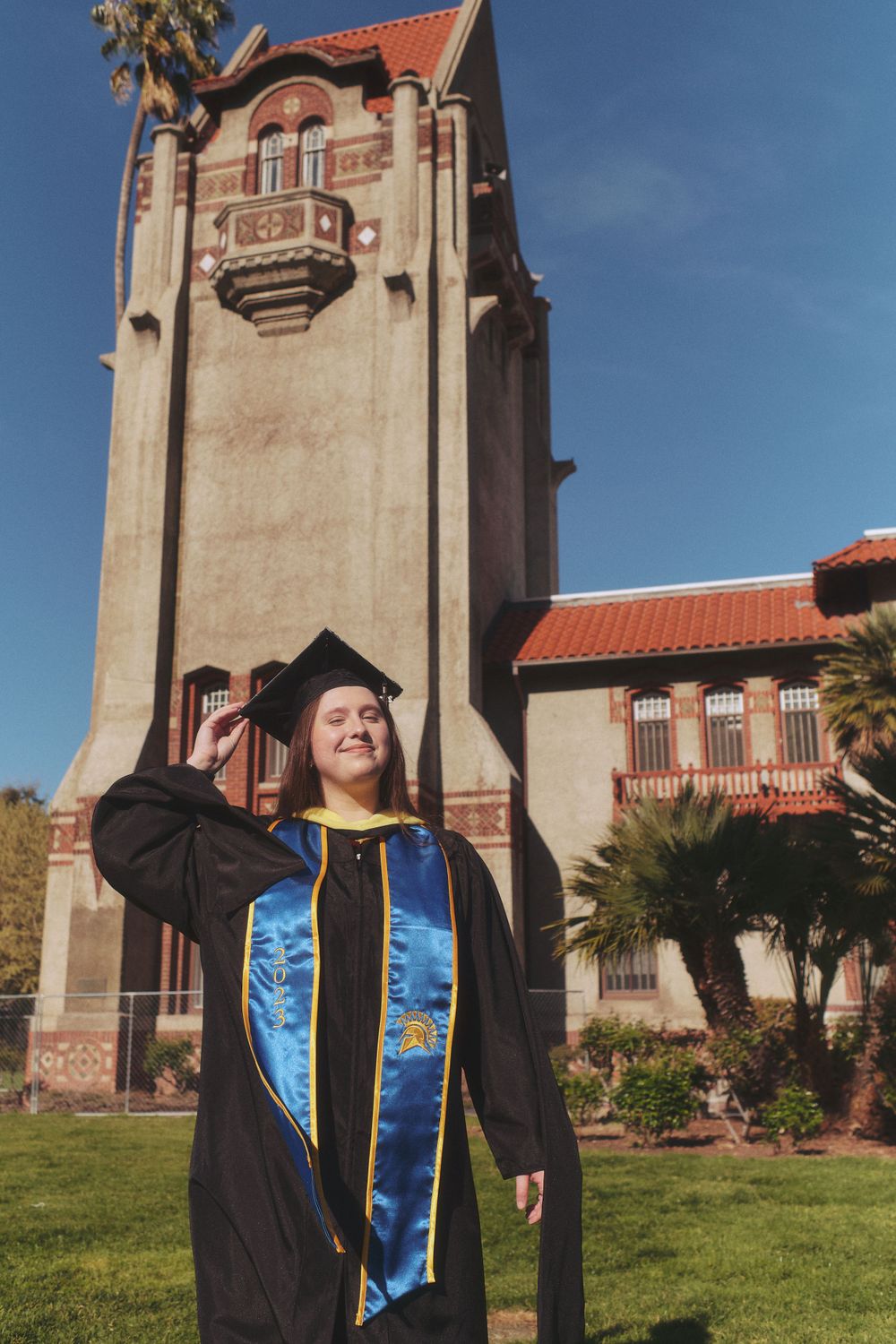 A graduate in black cap and gown with blue and gold stole stands before a historic campus tower.