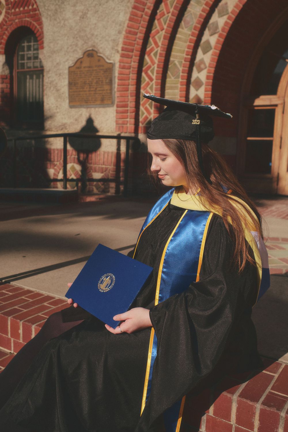 A graduate in cap and gown sits reviewing her blue diploma outside a red brick university building.