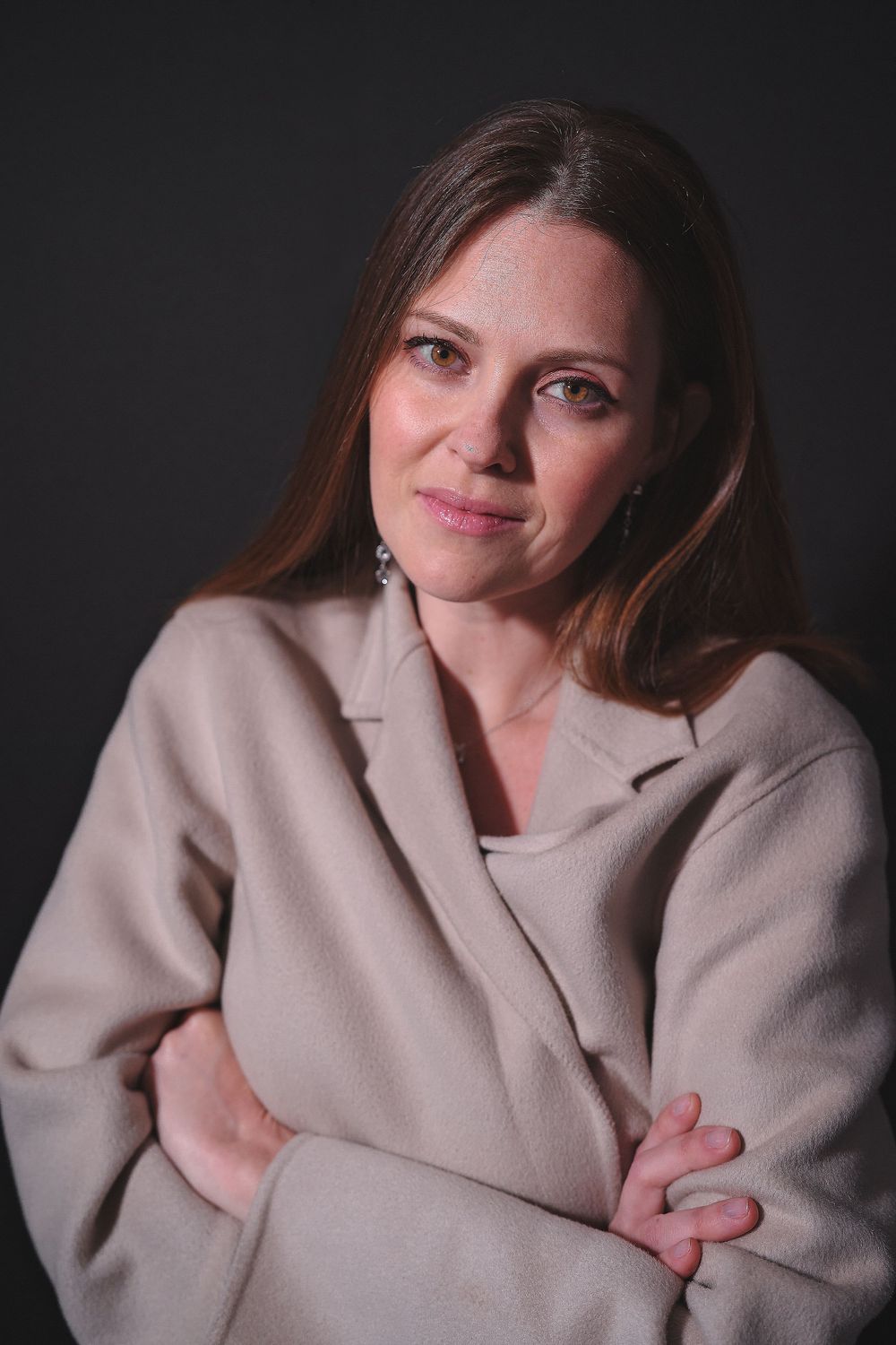 A brunette woman in a beige blazer smiles warmly with arms crossed against a dark background.