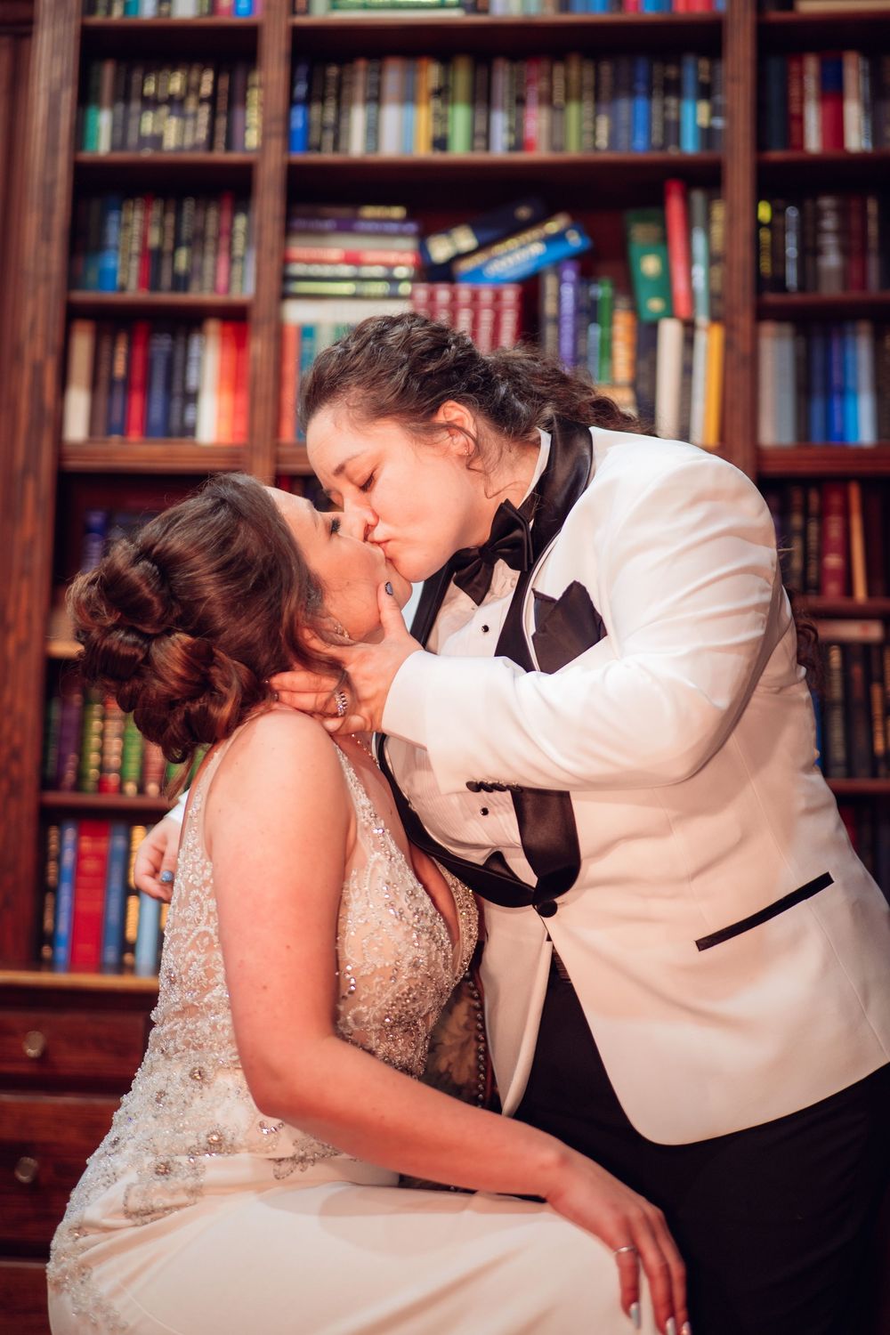 Couple shares intimate moments in a library setting with bookshelves in the background.