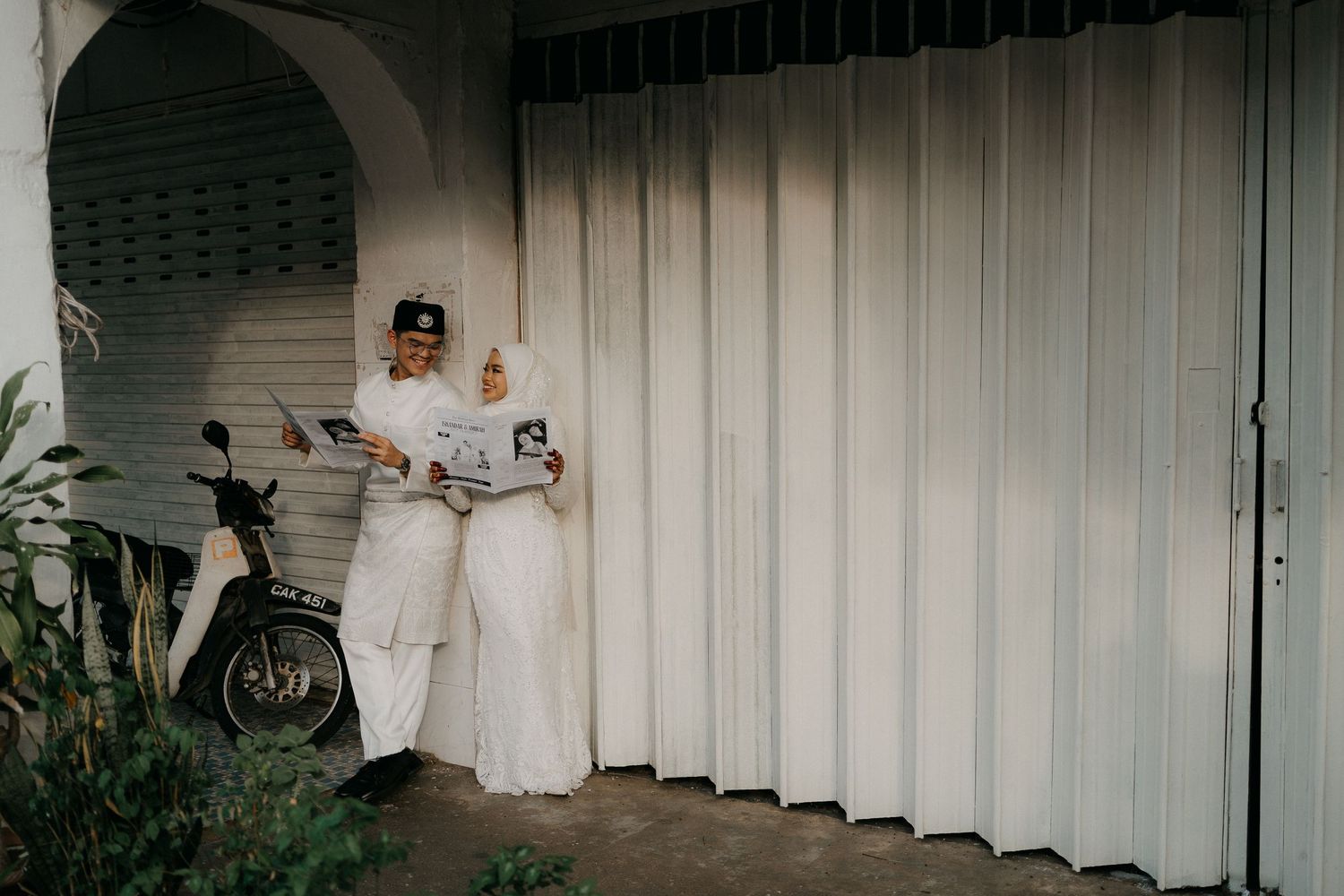 Malay bride and groom in traditional white wedding attire sharing a newspaper outside a vintage house in Malaysia. By Hizashi Studio