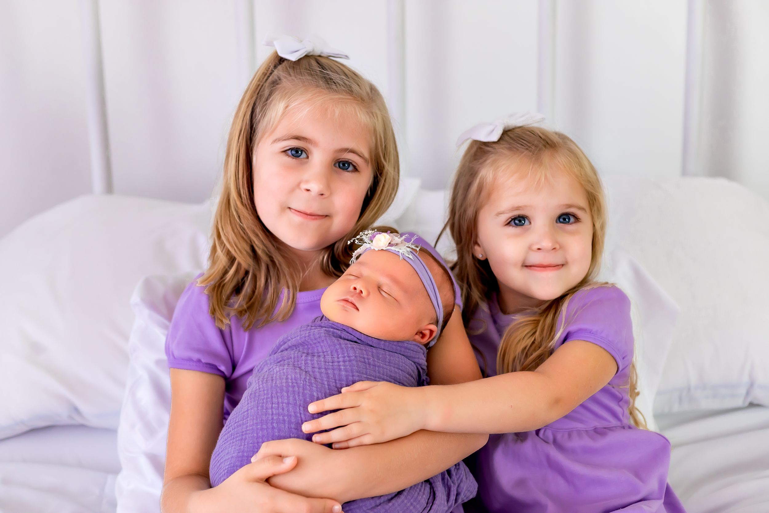 Three siblings in purple outfits together on white bedding.